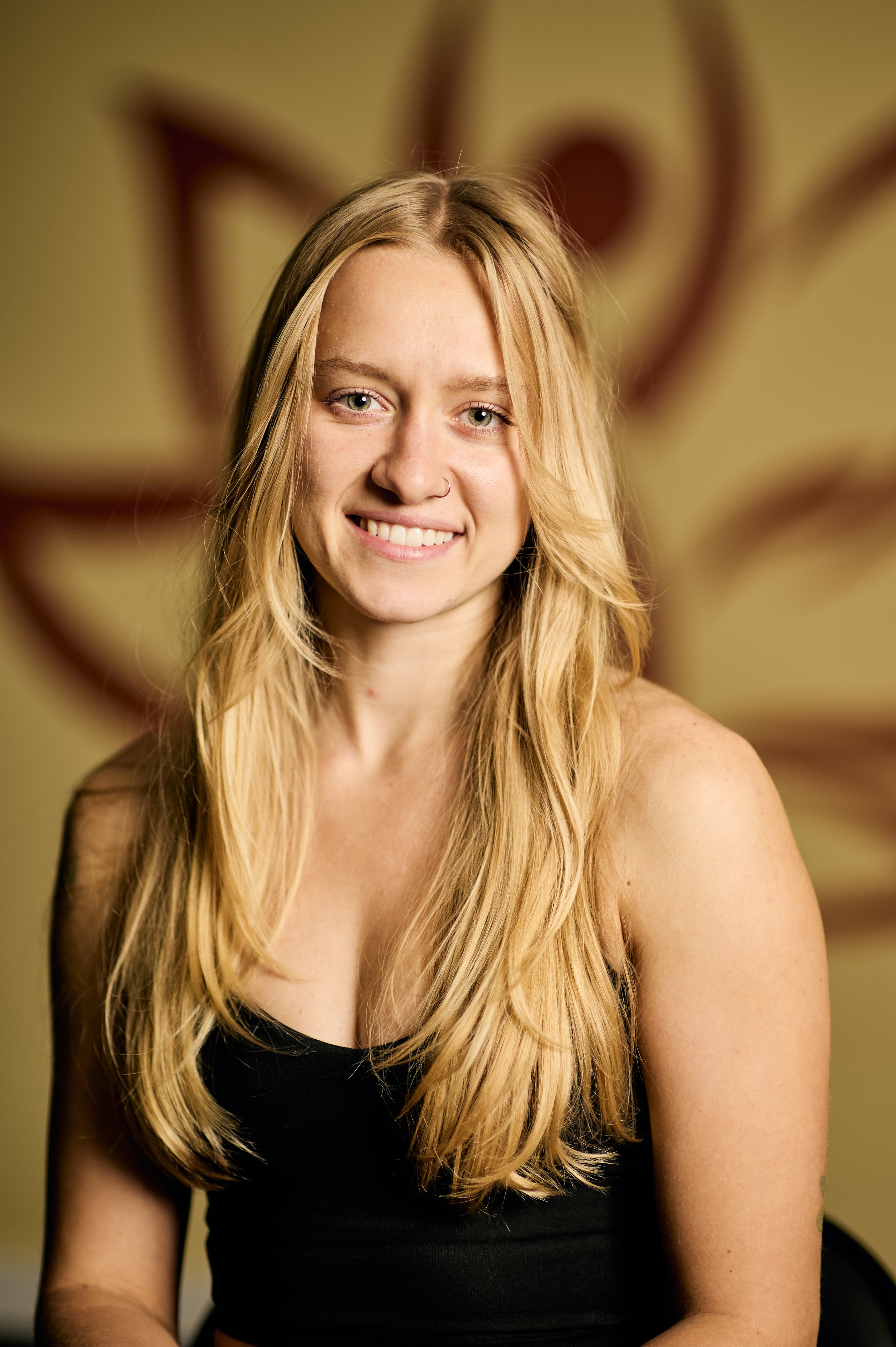 A smiling young woman with long blonde hair wearing a black sleeveless top, sitting indoors with a blurred decorative background.