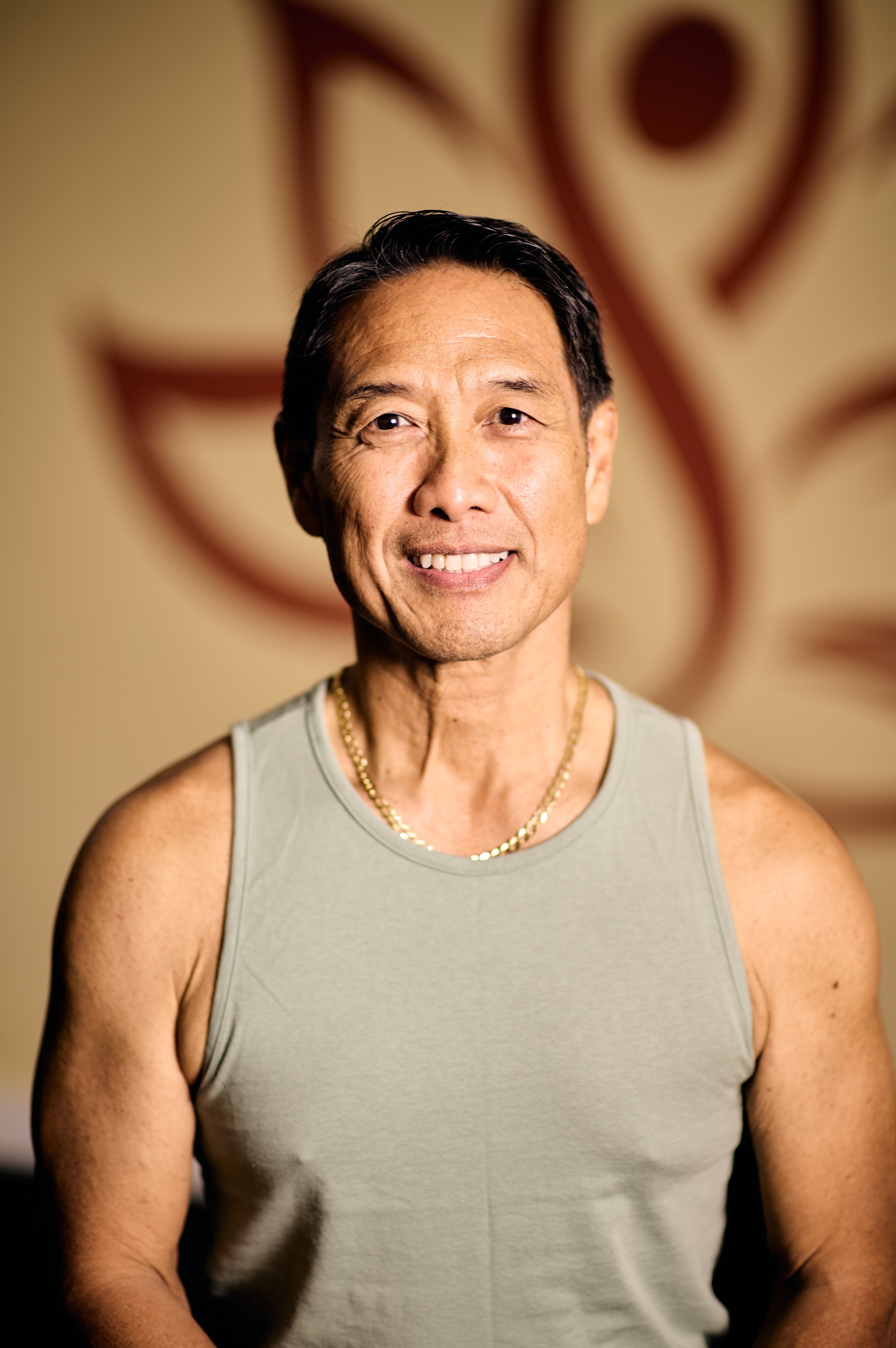 A middle-aged Asian man with short black hair, smiling, wearing a gray sleeveless shirt and a gold necklace, standing in front of a background with abstract art.