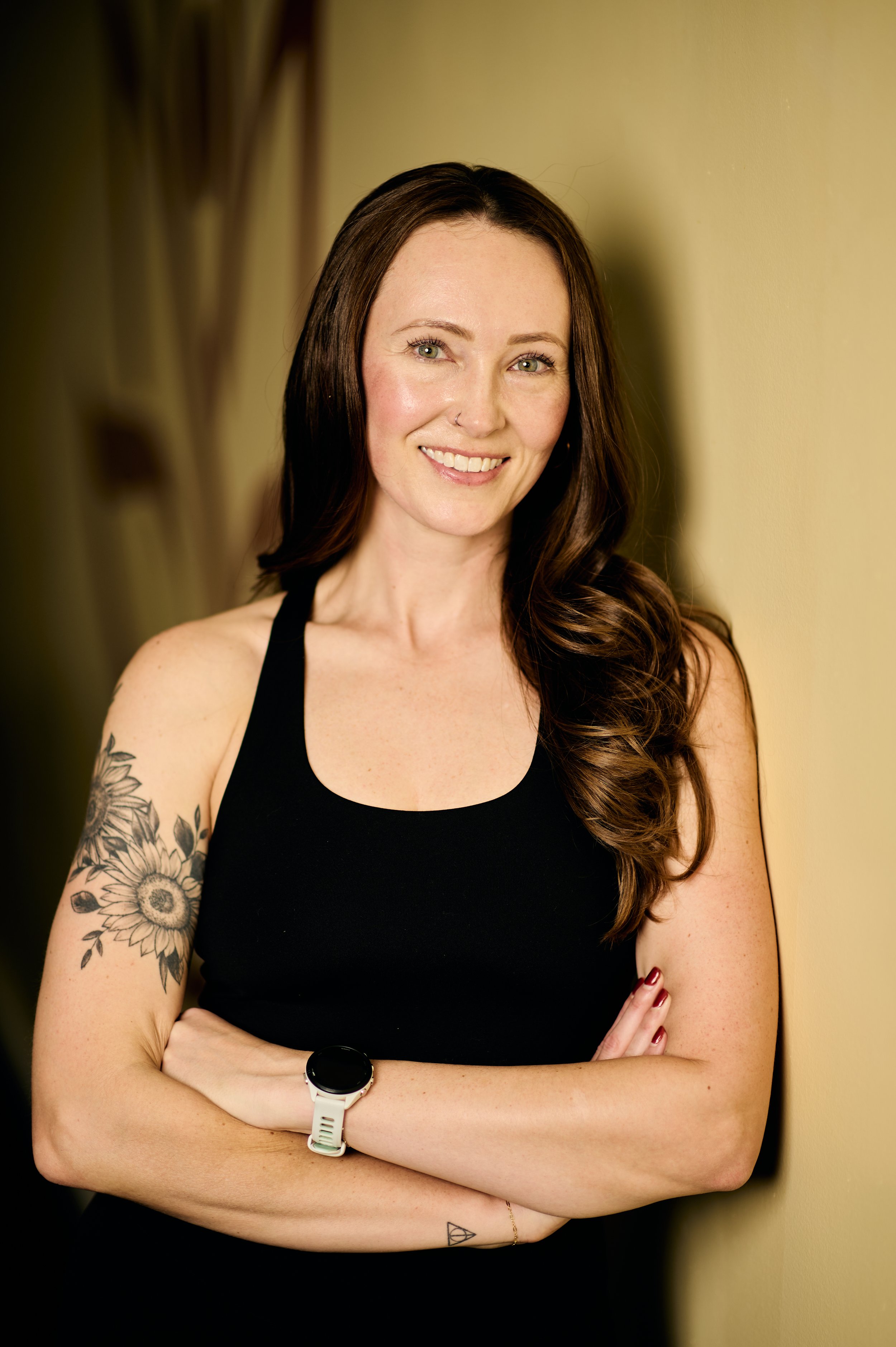 A woman with long brown hair and tattoos on her arm smiling while standing against a yellow wall.