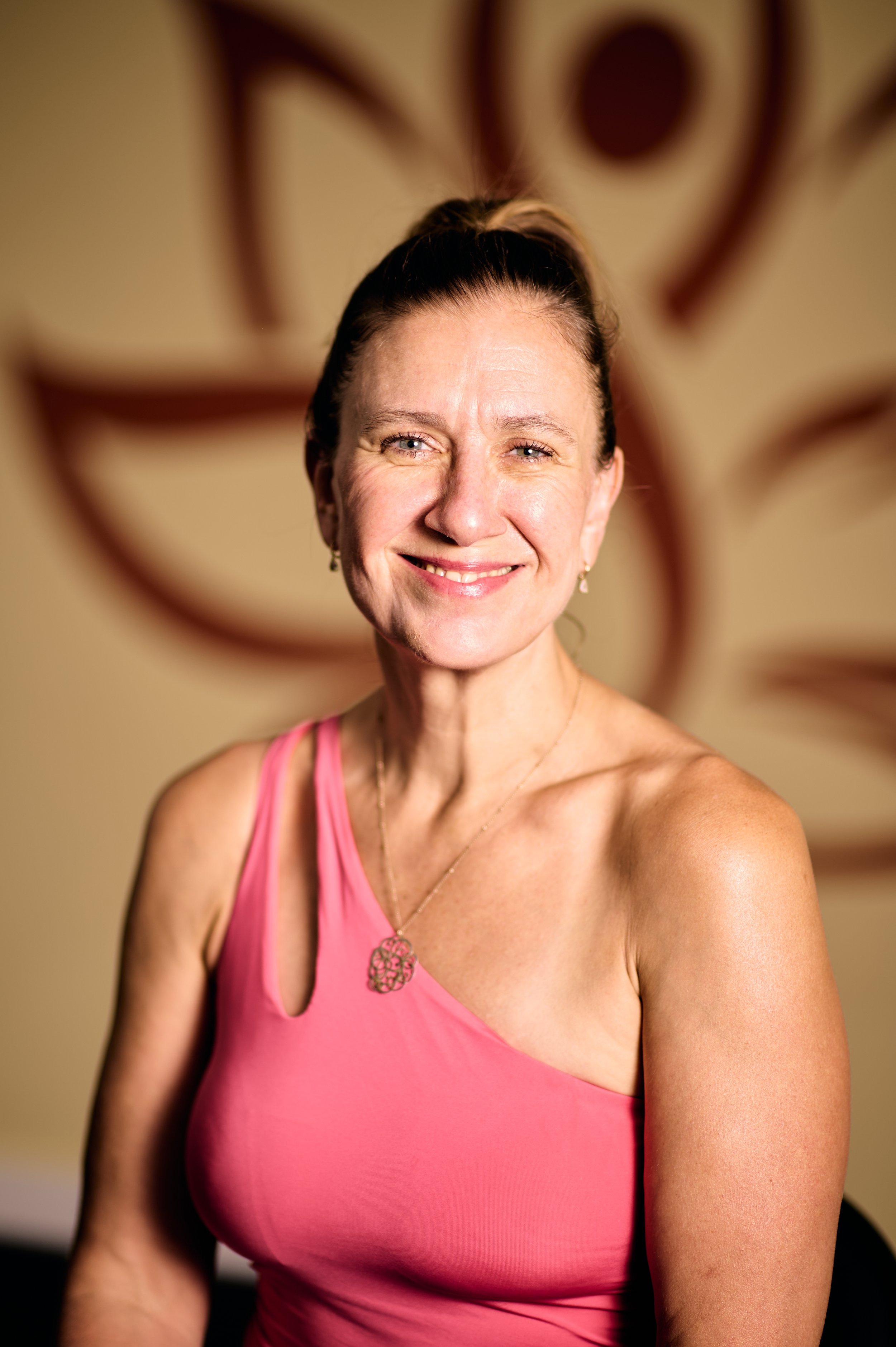 A smiling woman with dark hair in a ponytail, wearing a pink one-shoulder top and jewelry, in front of a beige background with a decorative pattern.
