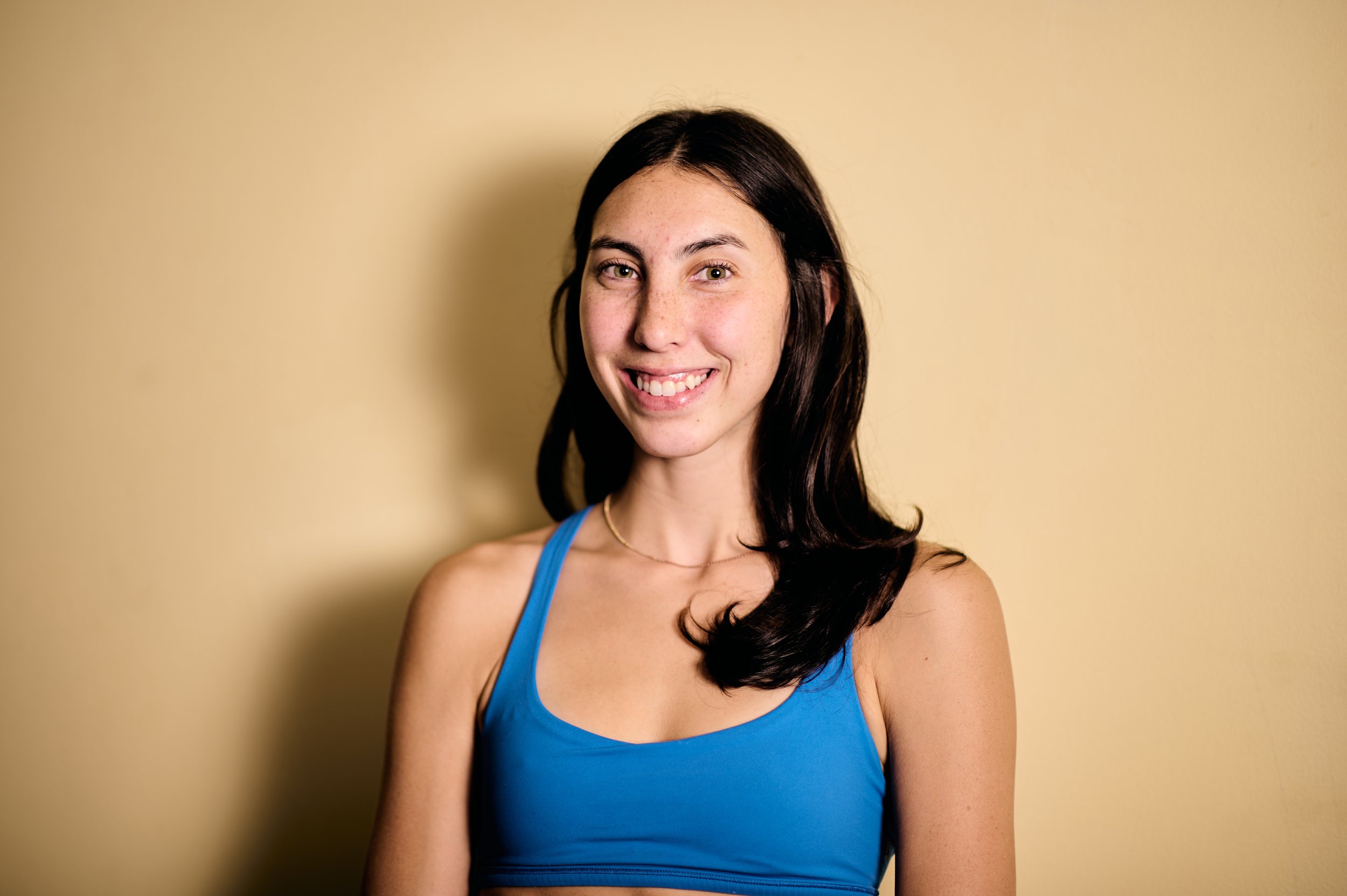 A young woman with long dark hair wearing a blue athletic tank top, smiling at the camera, standing against a plain beige wall.
