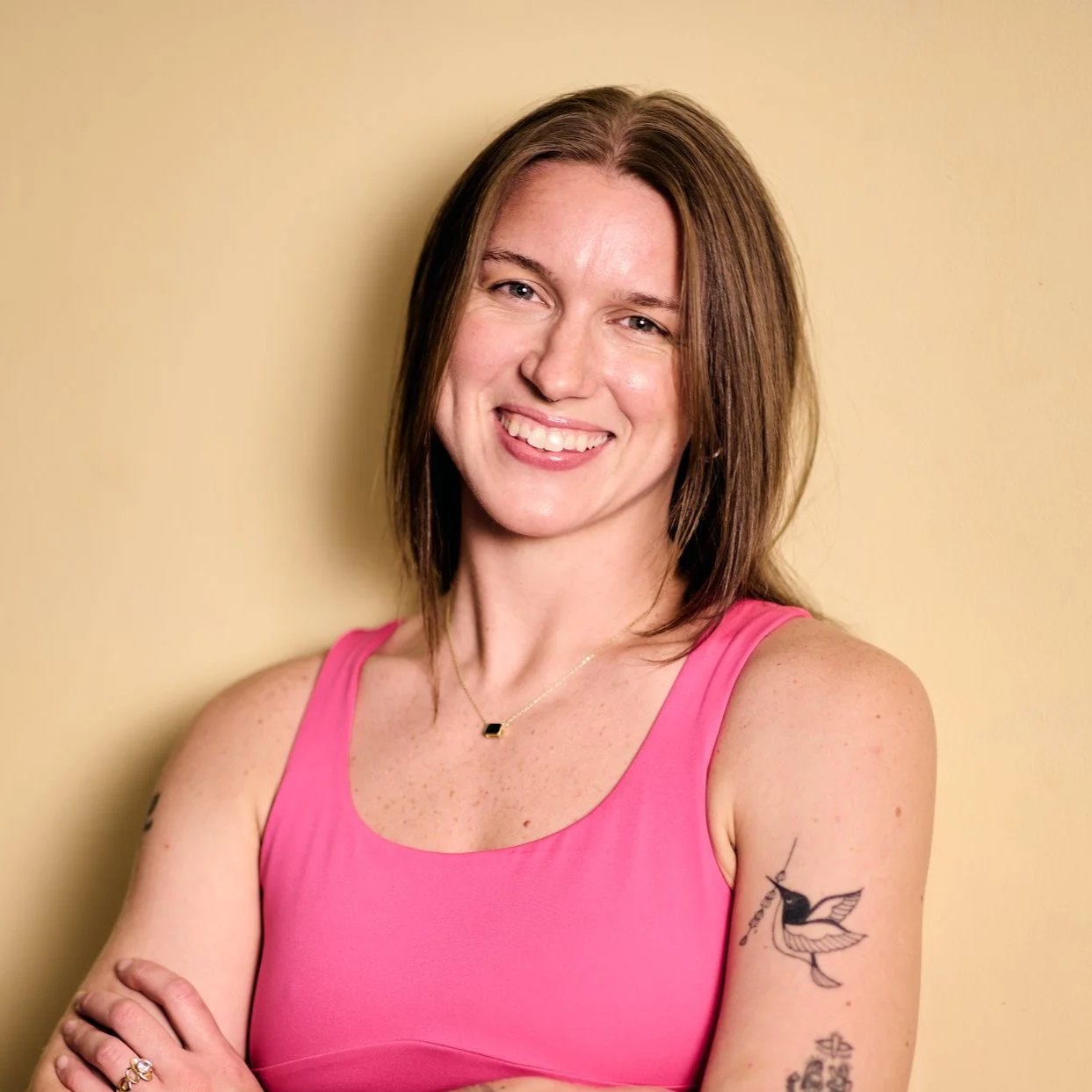 A woman with shoulder-length brown hair smiling, wearing a pink sleeveless top, standing against a beige background.