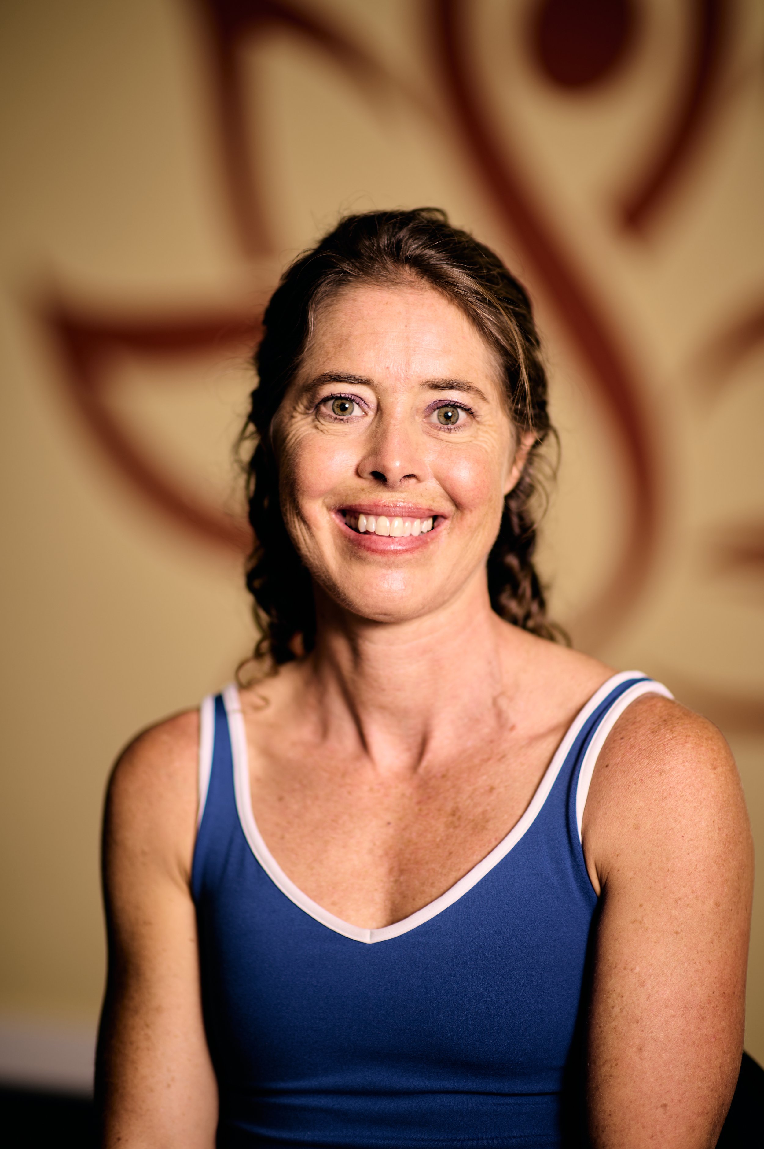 A woman with curly brown hair and green eyes smiling, wearing a blue tank top with white trim, in front of a blurred warm-toned background.