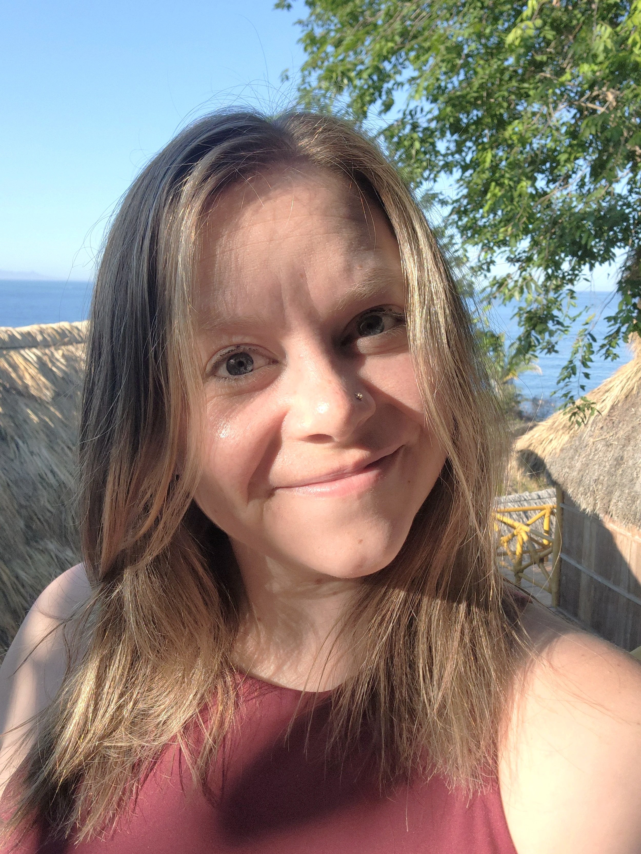 A young woman smiling outdoors near the ocean with a thatched roof structure and green trees in the background.