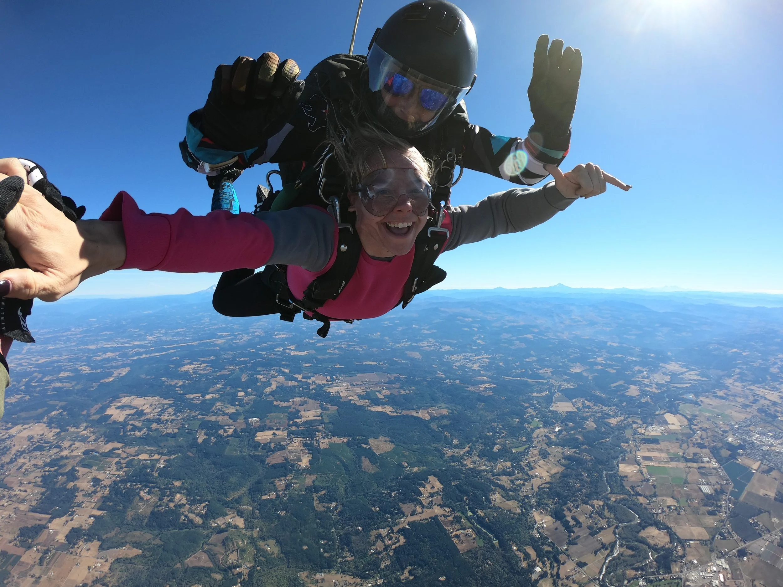 Two skydivers, a woman in pink and a woman in black helmet, are free-falling through the sky with a view of the landscape below, including fields, lakes, and distant mountains.