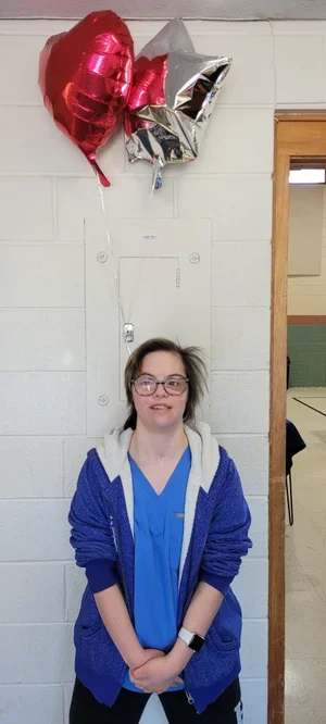 A girl with glasses and a blue hoodie standing in front of a white brick wall, holding her hands together. Three helium balloons—one red, one silver, and one gold—are floating above her.