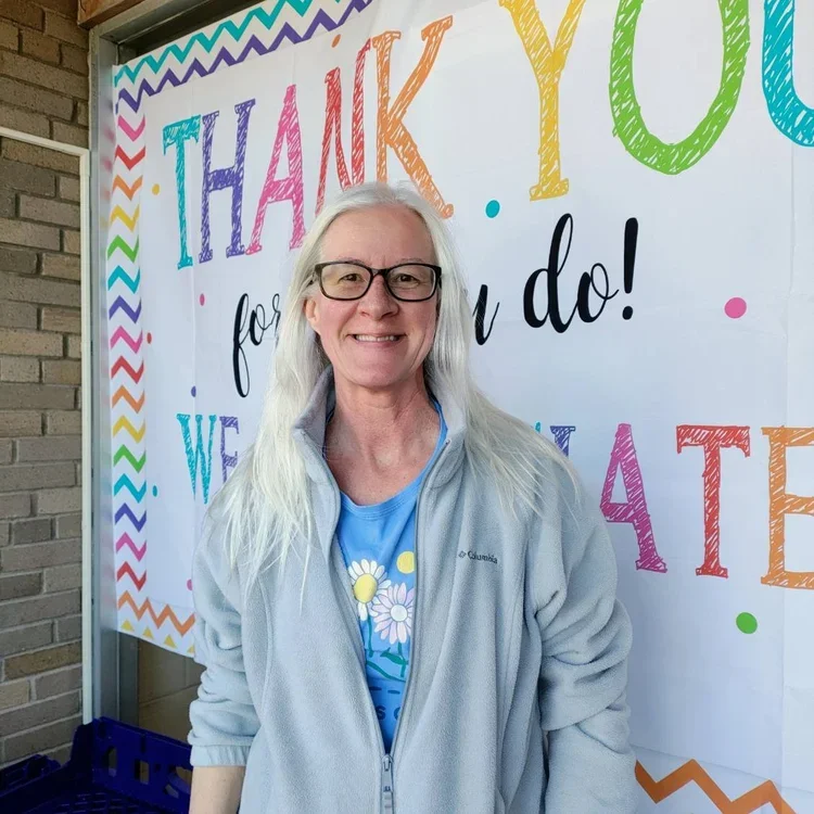 A woman with long white hair and glasses smiling in front of a colorful thank you banner.