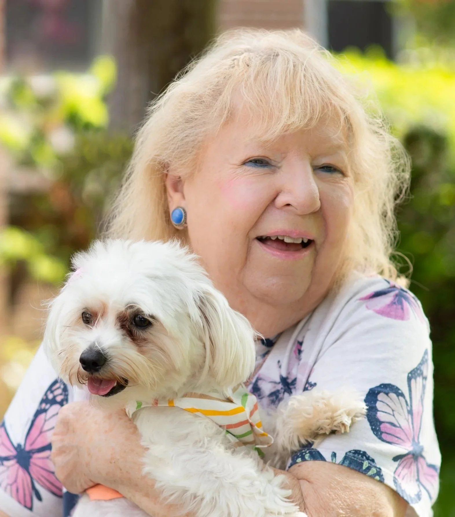 A smiling elderly woman with blonde hair wearing blue earrings, holding a small white dog with curly fur and a colorful striped bandana, outdoors with greenery in the background.