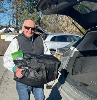A man wearing sunglasses and a black jacket stands next to an open car trunk, holding a large black package. There are parked cars and trees in the background.