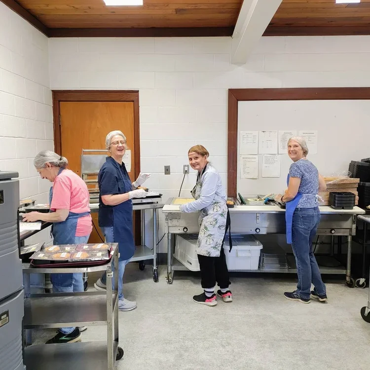 Four women working together in a kitchen or food preparation room, some wearing aprons and gloves, smiling and laughing.