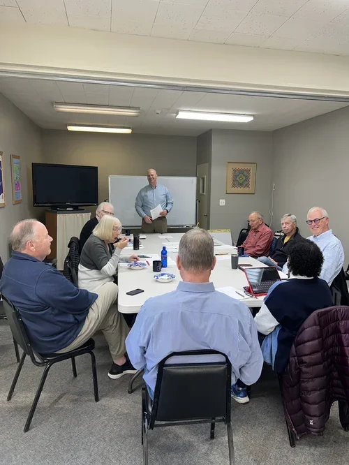 A man standing in front of a whiteboard leading a meeting with eight seated adults in a conference room.