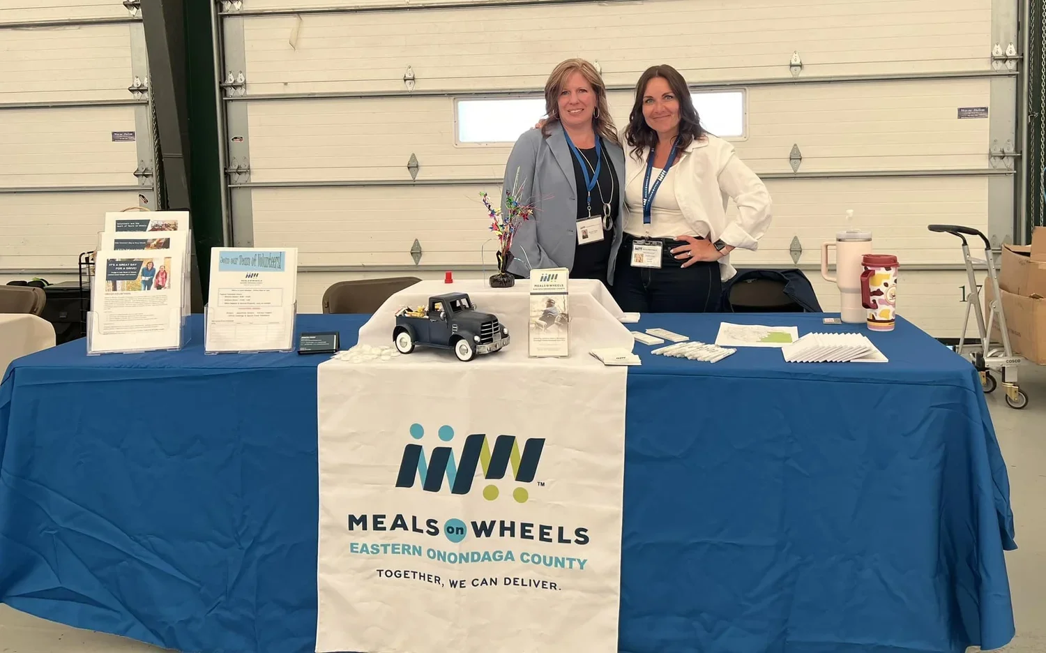 Two women standing behind a long table with a blue tablecloth and a sign that reads 'Meals on Wheels Eastern Onondaga County,' inside a garage or warehouse space. The table has brochures, a small decorative truck, and other informational materials.