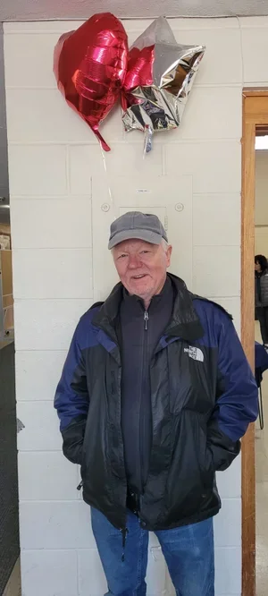 An elderly man wearing a gray baseball cap, black jacket, and blue jeans standing in front of balloons, including a red heart-shaped balloon, a metallic gray balloon, and a metallic gold balloon, attached to a white wall.