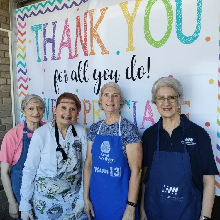 Four women wearing aprons and hairnets stand in front of a colorful thank you banner, smiling at the camera.