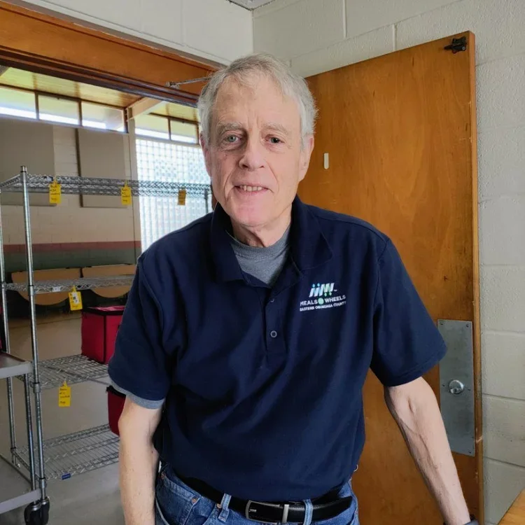 Older man with gray hair wearing a navy blue polo shirt and jeans standing indoors near a wooden door and metal shelving in a warehouse or storage room.