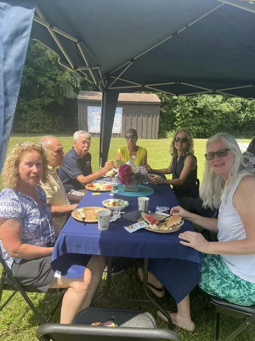 A group of six people sitting around a table outdoors under a canopy at a picnic or gathering. They are enjoying food and drinks on a sunny day with a grassy background, trees, and a small building.