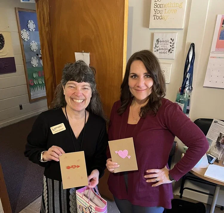Two women standing indoors, smiling and holding handmade cards with pink accents. One woman wears black with a name tag, the other wears a burgundy sweater.