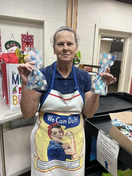 A woman wearing a Rosie the Riveter apron indoors, holding small gift bags, with holiday decorations in the background.