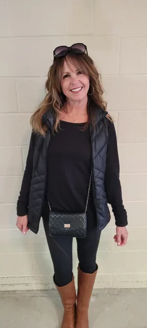 Woman with sunglasses on her head, wearing a black jacket, black top, and brown boots, standing against a beige wall.