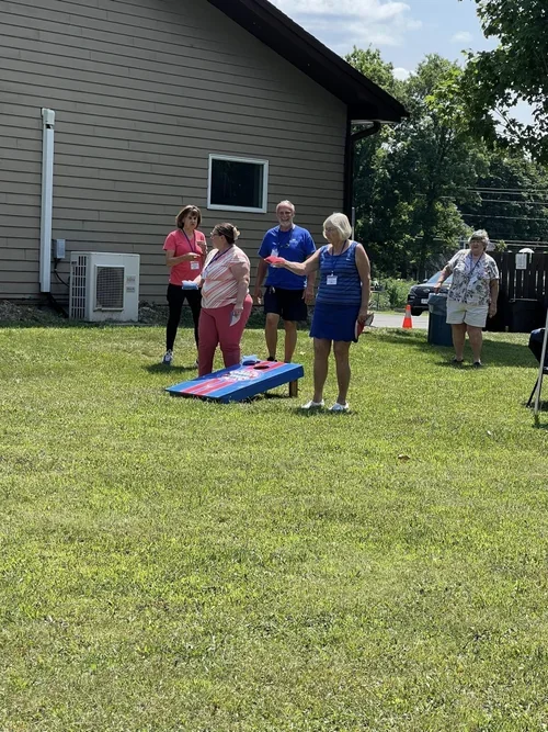 Group of five women and one man playing cornhole outdoors on a grassy lawn near a house.