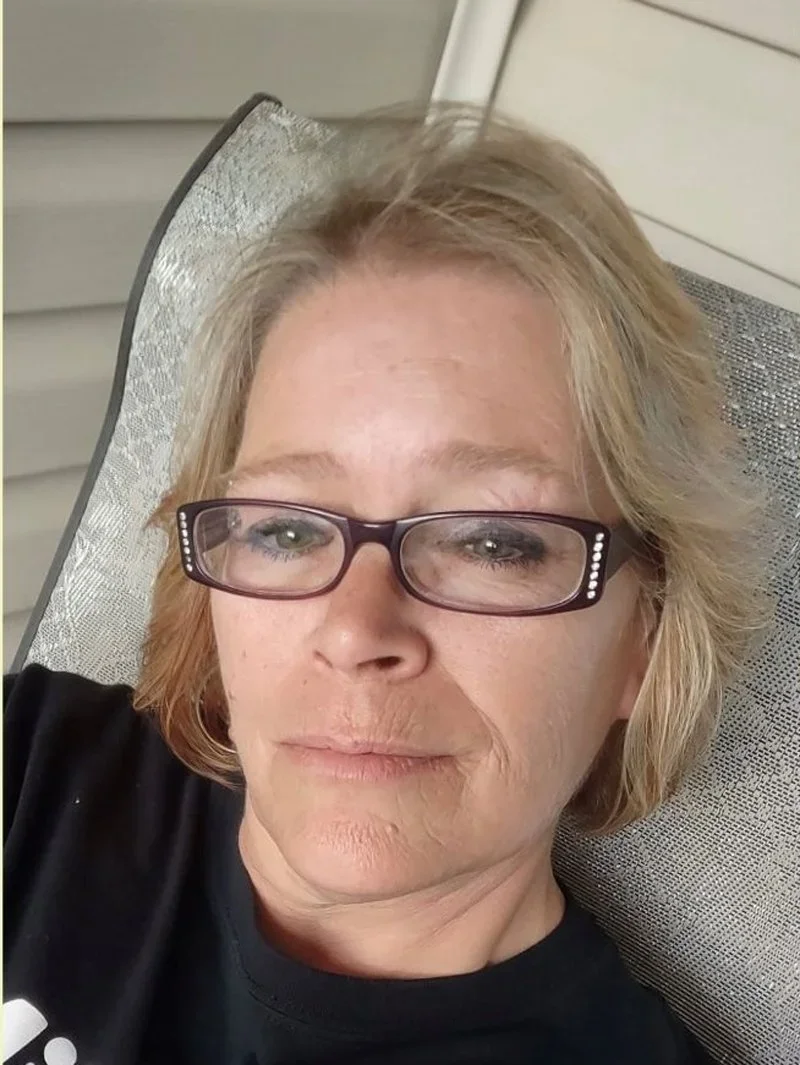 A woman with short blonde hair wearing glasses, lying on a patio chair with a water sprinkler overhead.