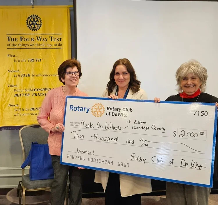 Three women standing together holding a large check for $2000 donor to Meals On Wheels, at a Rotary Club event in DeWitt. In the background, a yellow banner with the Rotary logo and 'The Four-Way Test' is visible.