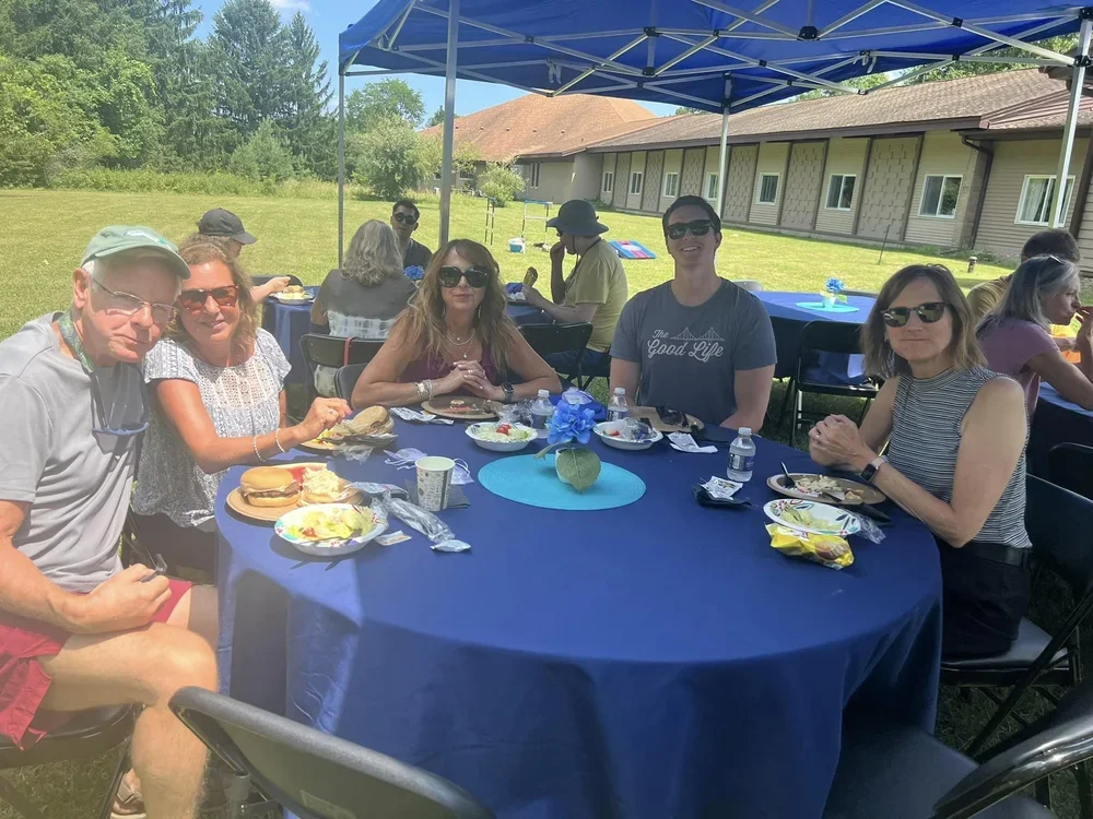 People sitting around a table outdoors during a meal, with more people in the background under a blue canopy, on a grassy area near a building.