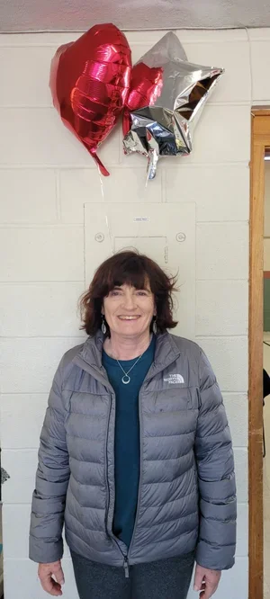 A woman standing indoors in front of a light-colored wall with three helium balloons in red, silver, and black shaped like hearts and stars, and smiling.