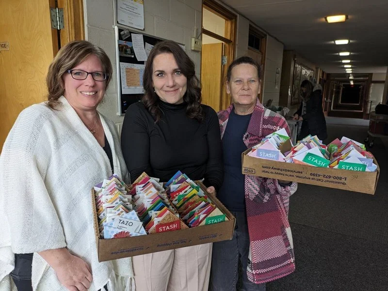 Three women standing indoors holding boxes filled with assorted colorful cards or papers.