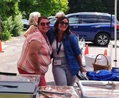 Three women smiling and posing together outdoors near a table with baked goods and a basket, with cars and trees in the background.