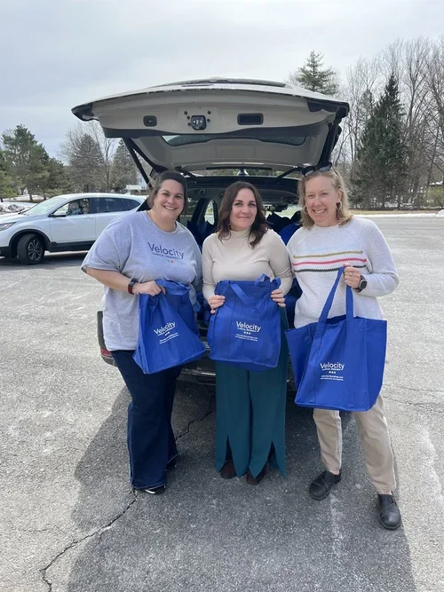 Three women standing in front of an open trunk of a car, holding blue reusable bags with 'Velocity' written on them, outdoors in a parking lot on a cloudy day.