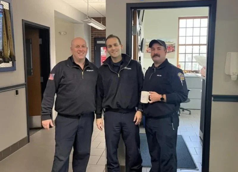 Three men in uniform standing together inside a building, smiling at the camera. One is holding a mug. They appear to be emergency or police personnel.
