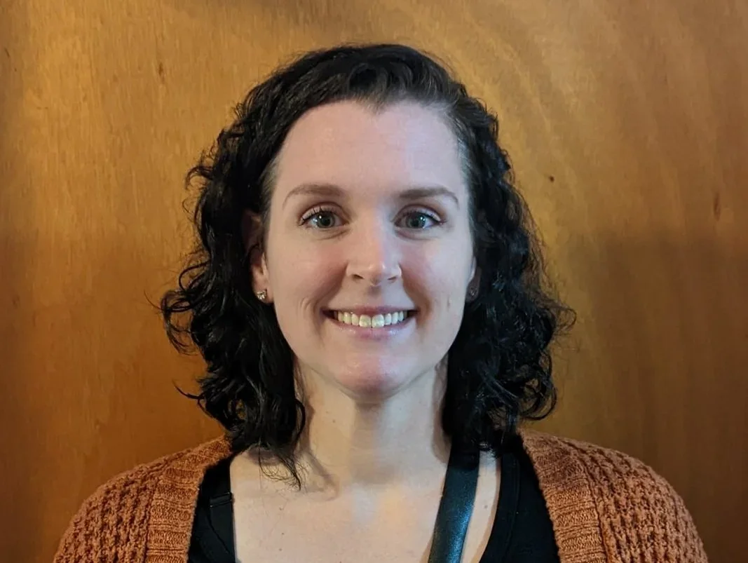A woman smiling, with dark curly hair, wearing a brown cardigan and a black top, standing against a wooden background.