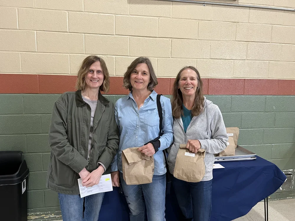 Three women smiling and holding paper bags at a table in an indoor setting with a beige and green brick wall background.