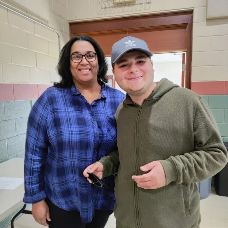 A woman with glasses and a blue plaid shirt standing next to a young man in a gray hoodie and baseball cap, both smiling indoors against a beige brick wall.