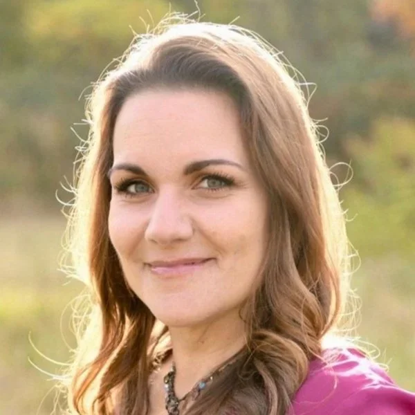 Close-up of a smiling woman with brown hair outdoors in natural light.