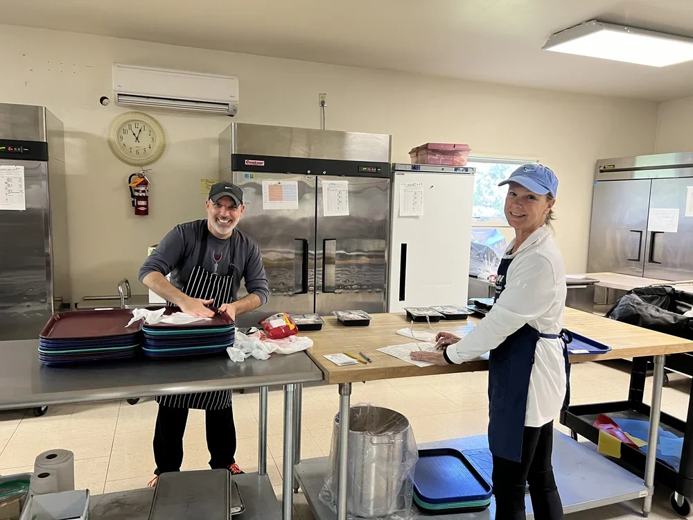 Two women in a commercial kitchen. One woman is smiling and wearing a gray cap and an apron, the other woman is smiling and wearing a blue cap and a white apron. They are standing at a work table with trays, papers, and utensils. Behind them are larg
