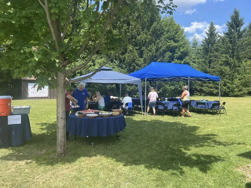 Outdoor gathering with tables and tents set up on a grassy area, with people serving food and sitting at tables under blue and gray canopies, surrounded by trees.