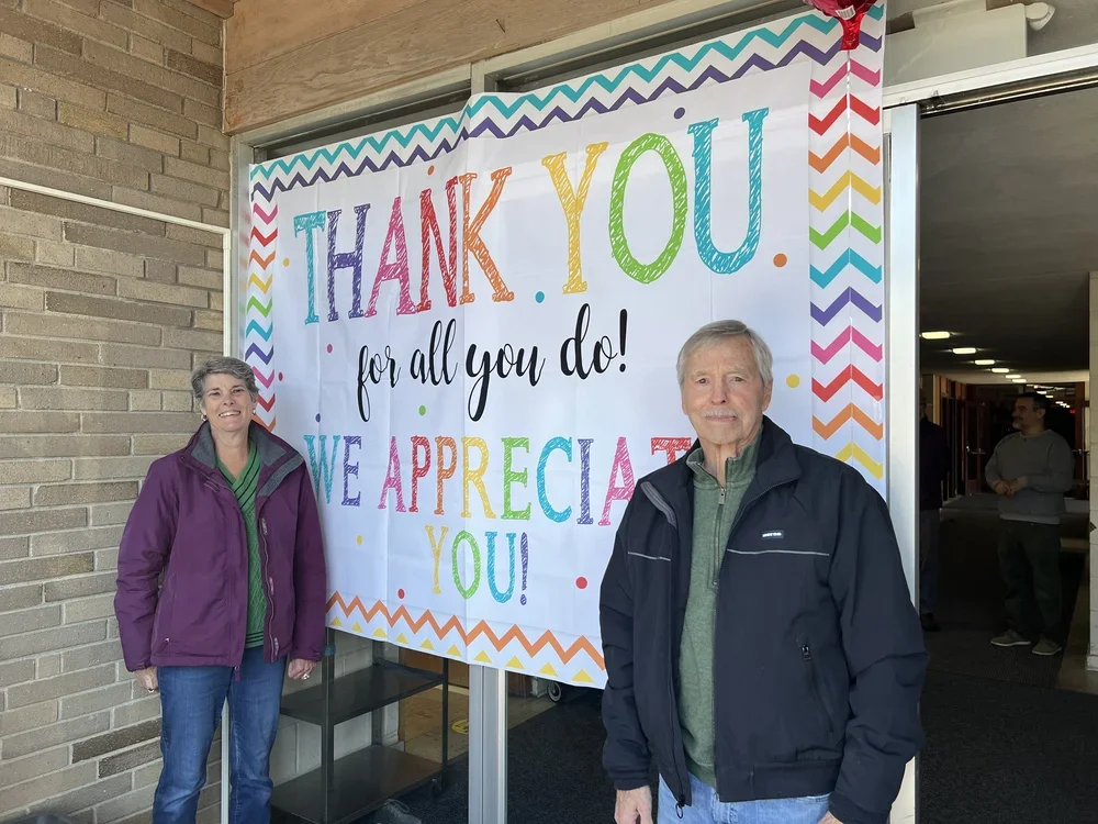Two people standing in front of a colorful thank you banner at a school or community center. The banner reads 'Thank you for all you do! We appreciate you!' in bright, multicolored letters.