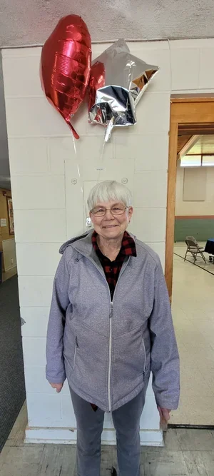 An elderly woman standing indoors with three helium balloons—red, silver, and star-shaped—floating above her.