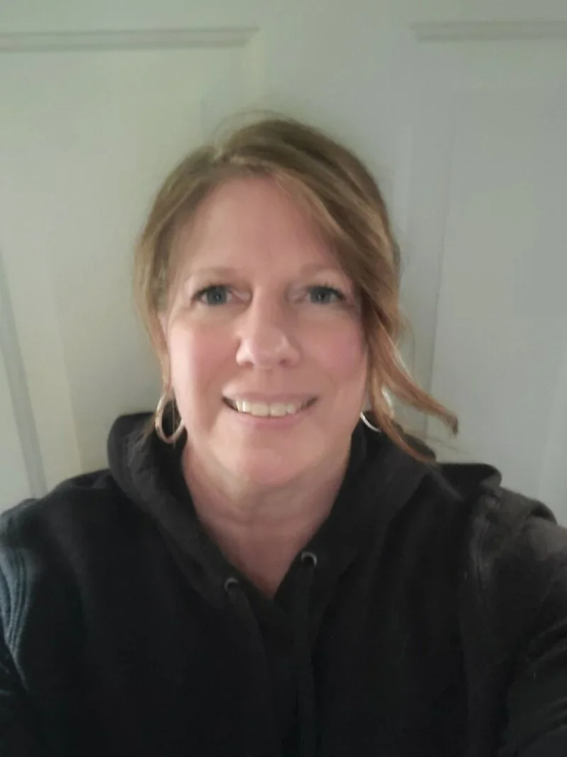 A woman with short, light brown hair smiling at the camera, wearing a black hoodie, standing in front of a light-colored door.