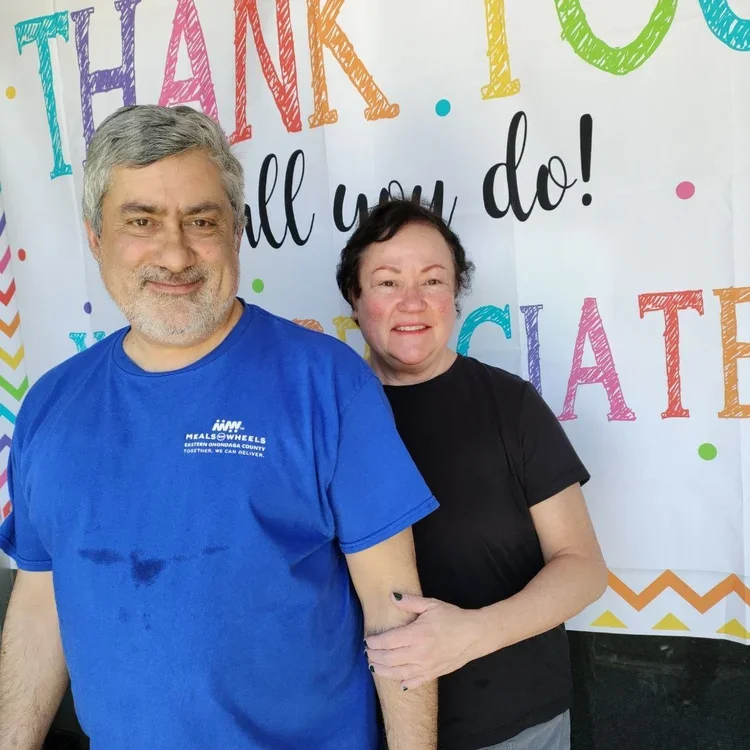 Two smiling people standing in front of a colorful thank you banner at a community event.