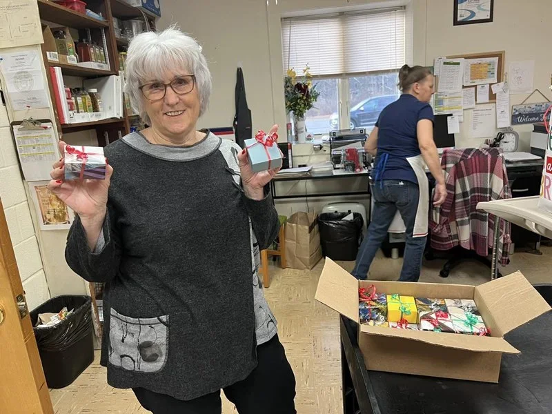 A smiling elderly woman with white hair and glasses holding up two small wrapped Christmas gifts inside a room with shelves and a window. In the background, another woman is working near a counter, and a large cardboard box filled with more wrapped p