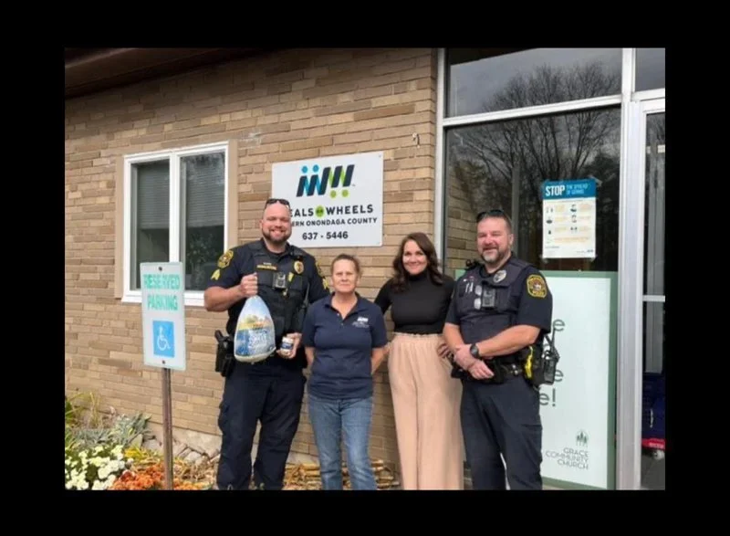 Two police officers and two women standing outside a building with a sign that reads 'Neul's Wheels' and 'Northern Onondaga County'. The police officers are smiling and holding items, and the group appears to be in front of a community or organizatio