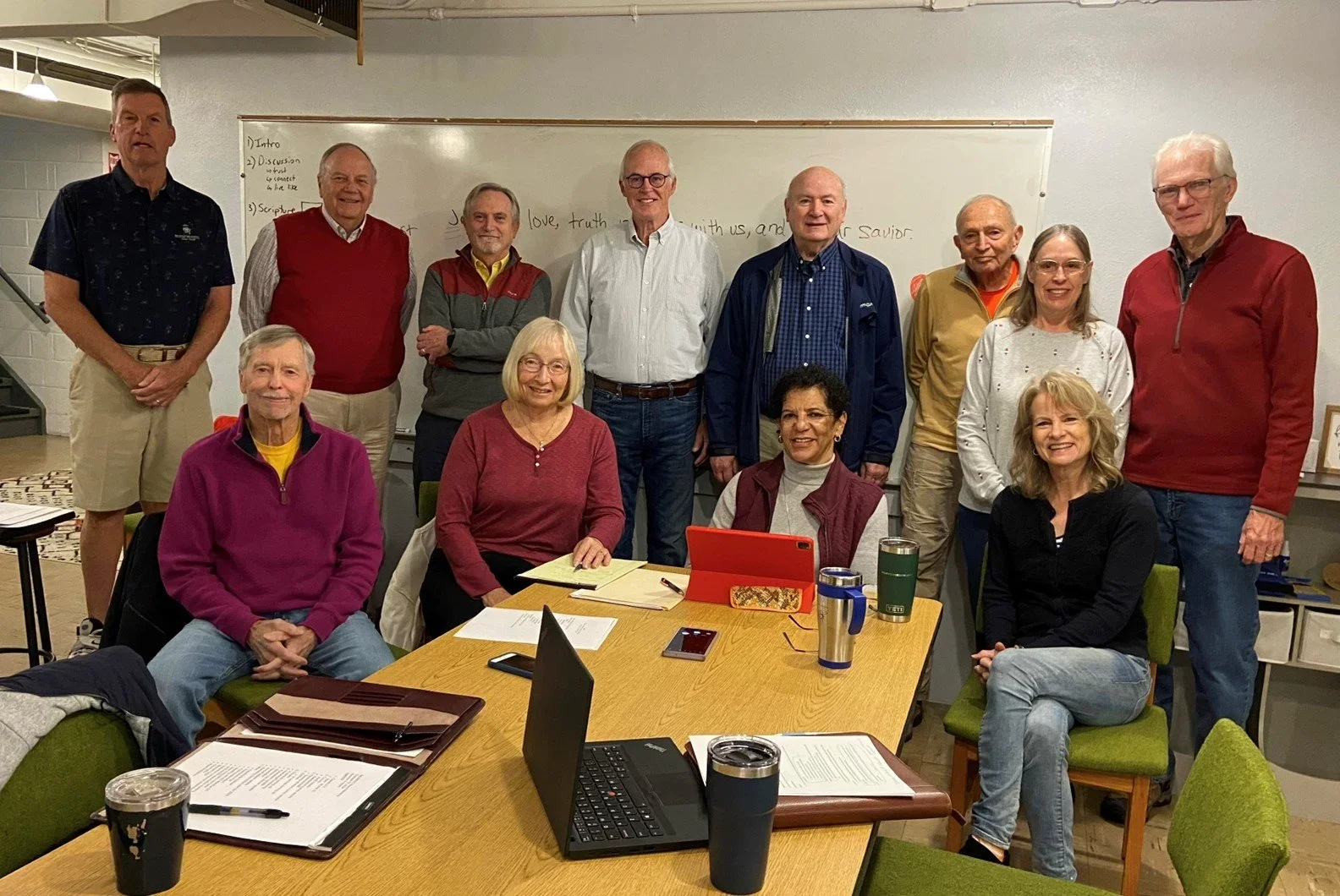 Group of fifteen diverse adults gathered in a conference room, some seated at a table with laptops and papers, others standing behind, smiling and posing for the photo.