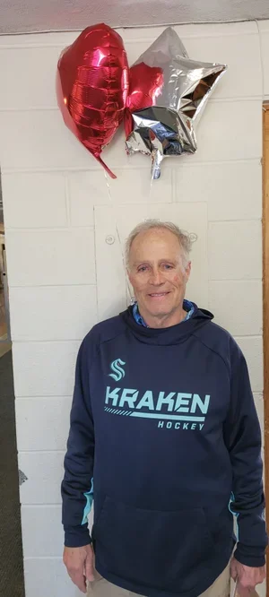 Older man standing in front of a white wall with three helium balloons (red heart-shaped, silver star-shaped, and gray star-shaped) attached to the wall above his head, wearing a blue Kraken hockey hoodie.