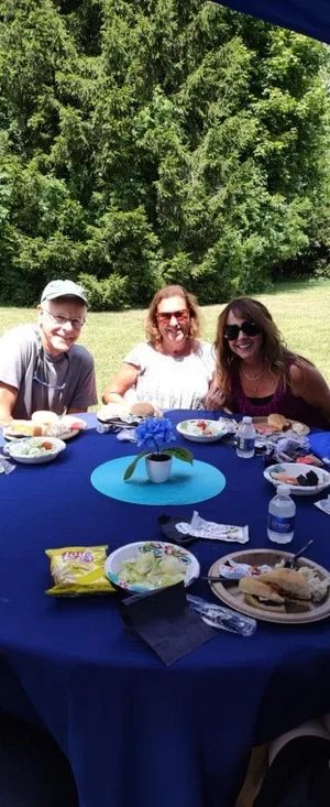 Three people sitting at a round table outdoors under a blue canopy, with food and drinks on the table, surrounded by green trees.