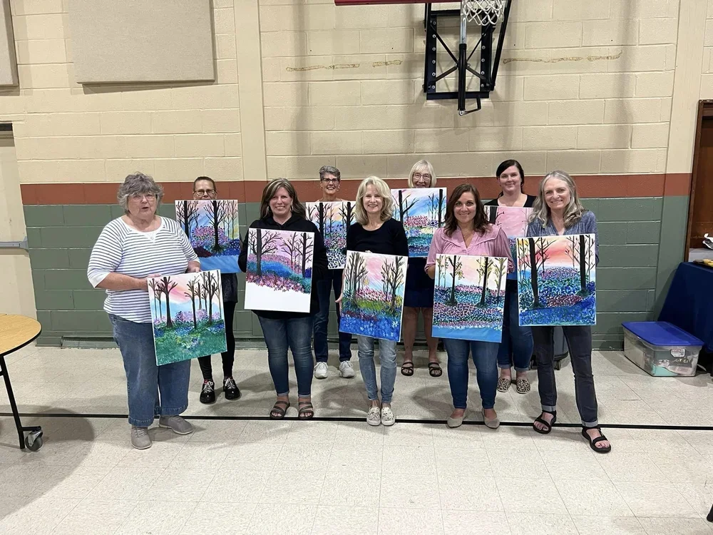 Group of nine women standing in a gymnasium holding colorful landscape paintings of trees at sunset or sunrise.