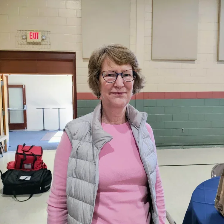 A woman with short brown hair, glasses, wearing a pink shirt and a gray vest, standing indoors in front of a beige brick wall with a red exit sign above a door.