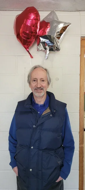 An older man with gray hair and a beard smiling, standing in front of a white wall with three helium balloons (a red heart, a silver star, and a gold star) floating above his head.